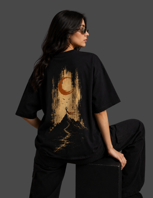 A woman wearing an oversized black t-shirt with a large graphic print on the back, featuring a crescent moon and mountain design in gold and brown tones, posed against a dark gray background.