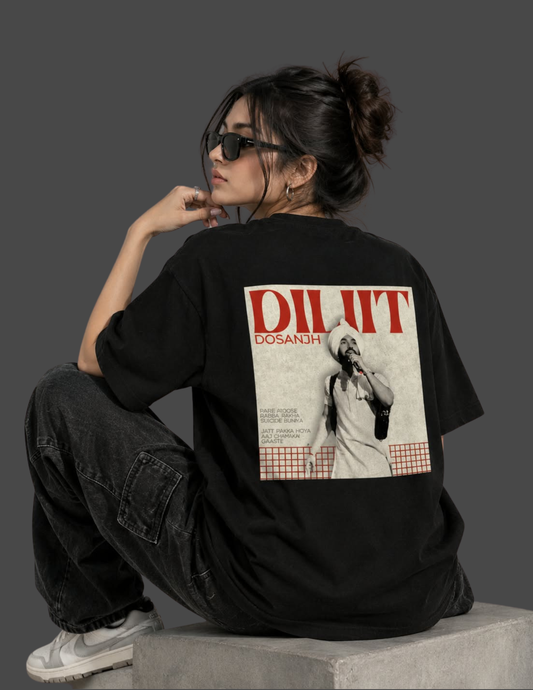 A woman wearing a black oversized t-shirt with a large graphic print on the back, sitting on a concrete block against a dark gray studio background.