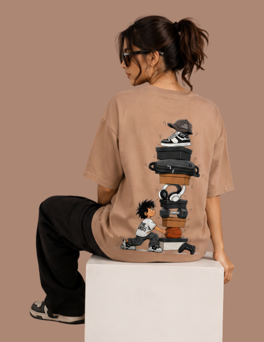 A woman wearing a beige oversized graphic t-shirt, shown from the back while sitting on a white block. The t-shirt features a detailed graphic of stacked sneakers, headphones, and a cartoon character.