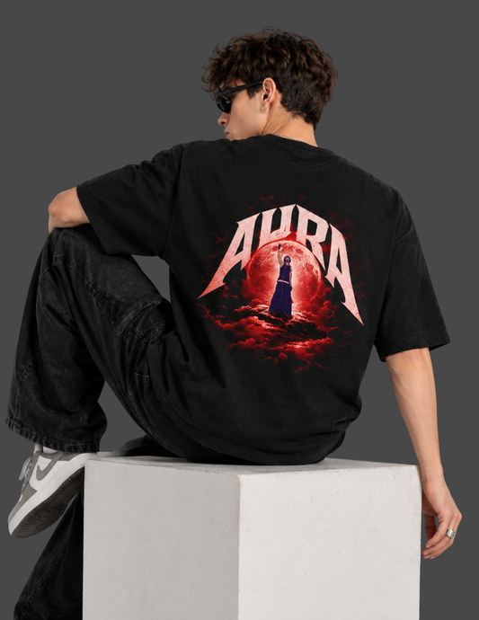 A man wearing a black t-shirt with a large red and white 'AURA' graphic on the back, sitting on a white block against a dark gray background.