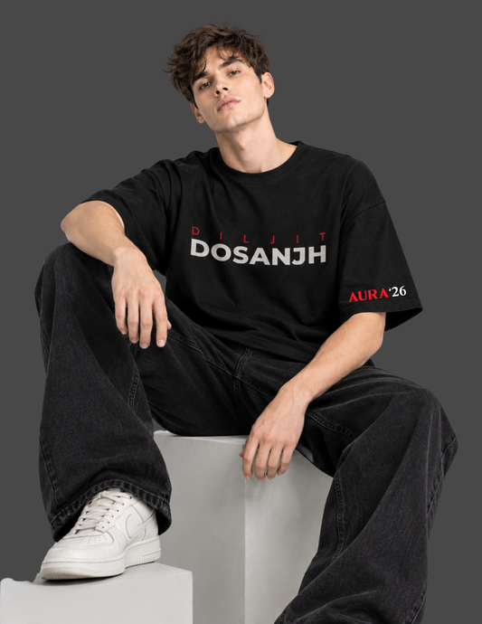 A man wearing a black Dosanjh t-shirt with 'DILJIT DOSANJH' printed in white and red on the chest, paired with black jeans and white sneakers, sitting on a white block against a dark gray background.