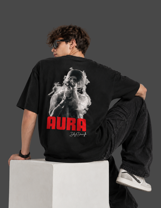 A model wearing a black graphic t-shirt with a large back print of a woman smoking and the word 'AURA' in red, sitting on a white block against a dark gray background.