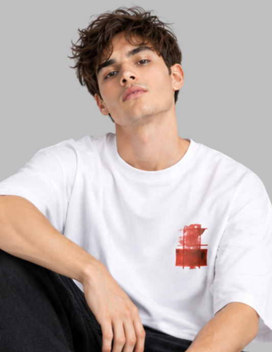 A young man with wavy brown hair wearing a white crewneck t-shirt with a red abstract graphic on the left chest, posed against a solid gray background.
