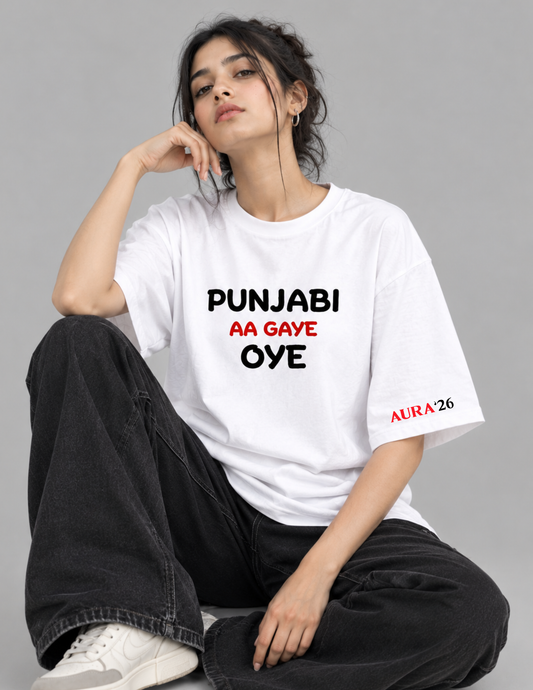 A woman sitting cross-legged wearing a white graphic t-shirt with the Punjabi text 'PUNJABI AA GAYE OYE' in black and red, paired with black jeans and white sneakers, against a solid gray studio background.
