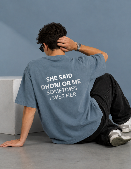A man sitting on a concrete floor, viewed from behind, wearing a faded blue t-shirt with white text that reads 'SHE SAID DHONI OR ME SOMETIMES I MISS HER'.