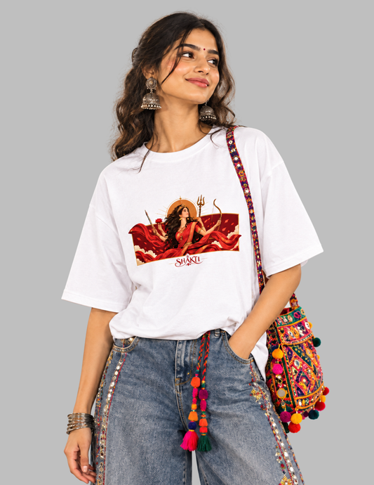 A woman wearing a white graphic t-shirt with a red and gold Diwali illustration, paired with blue jeans and a colorful embroidered shoulder bag, set against a solid gray background.
