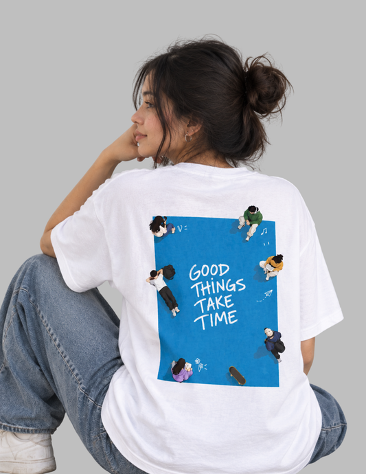 A woman wearing a white graphic t-shirt, viewed from behind. The shirt features a large blue rectangular graphic on the back with the text 'GOOD THINGS TAKE TIME' in white, surrounded by small cartoon illustrations of people.