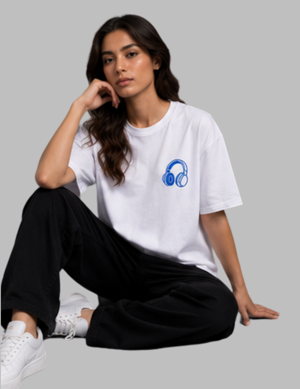A woman sitting cross-legged wearing a white crewneck t-shirt with a blue headphones graphic on the left chest, paired with black sweatpants and white sneakers, against a solid gray background.