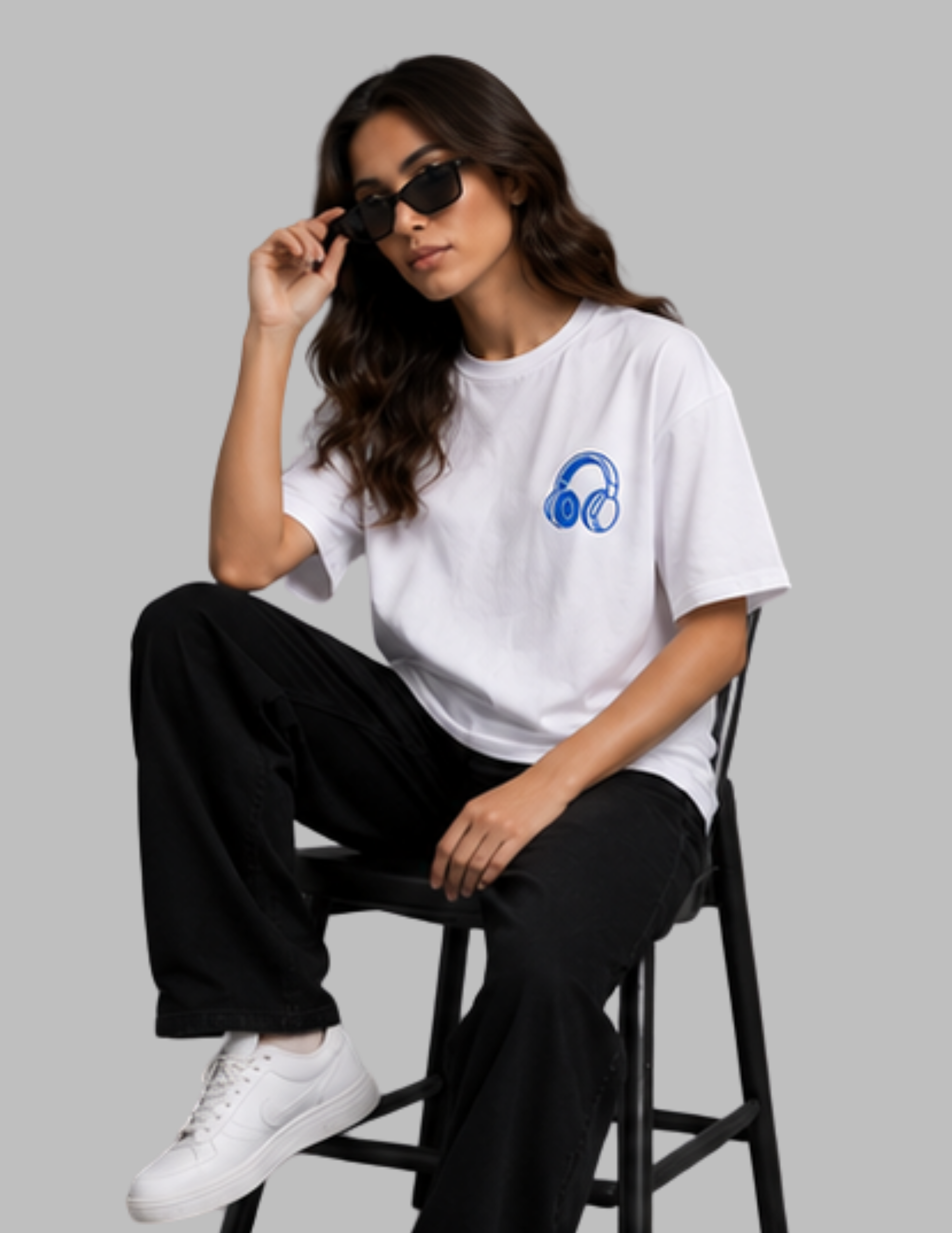 A woman sits on a black stool wearing a white short-sleeve T-shirt with a blue headphones graphic on the left chest, black pants, white sneakers, and dark sunglasses against a solid gray background.