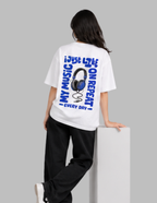 A model from behind wearing a white t-shirt with a large blue graphic print on the back, paired with black sweatpants, leaning on a white block against a gray background.