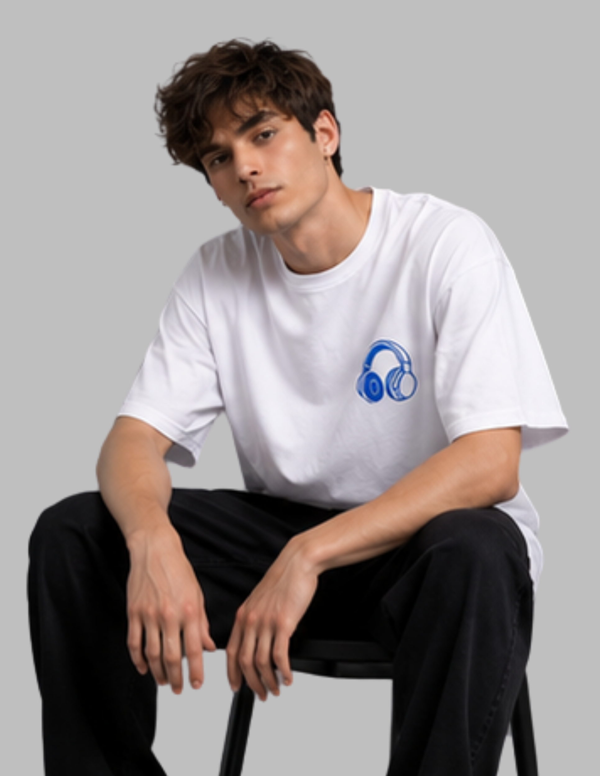 A young man sitting on a black stool wearing a white crewneck t-shirt with a blue headphones graphic on the left chest, paired with black pants, against a solid gray background.