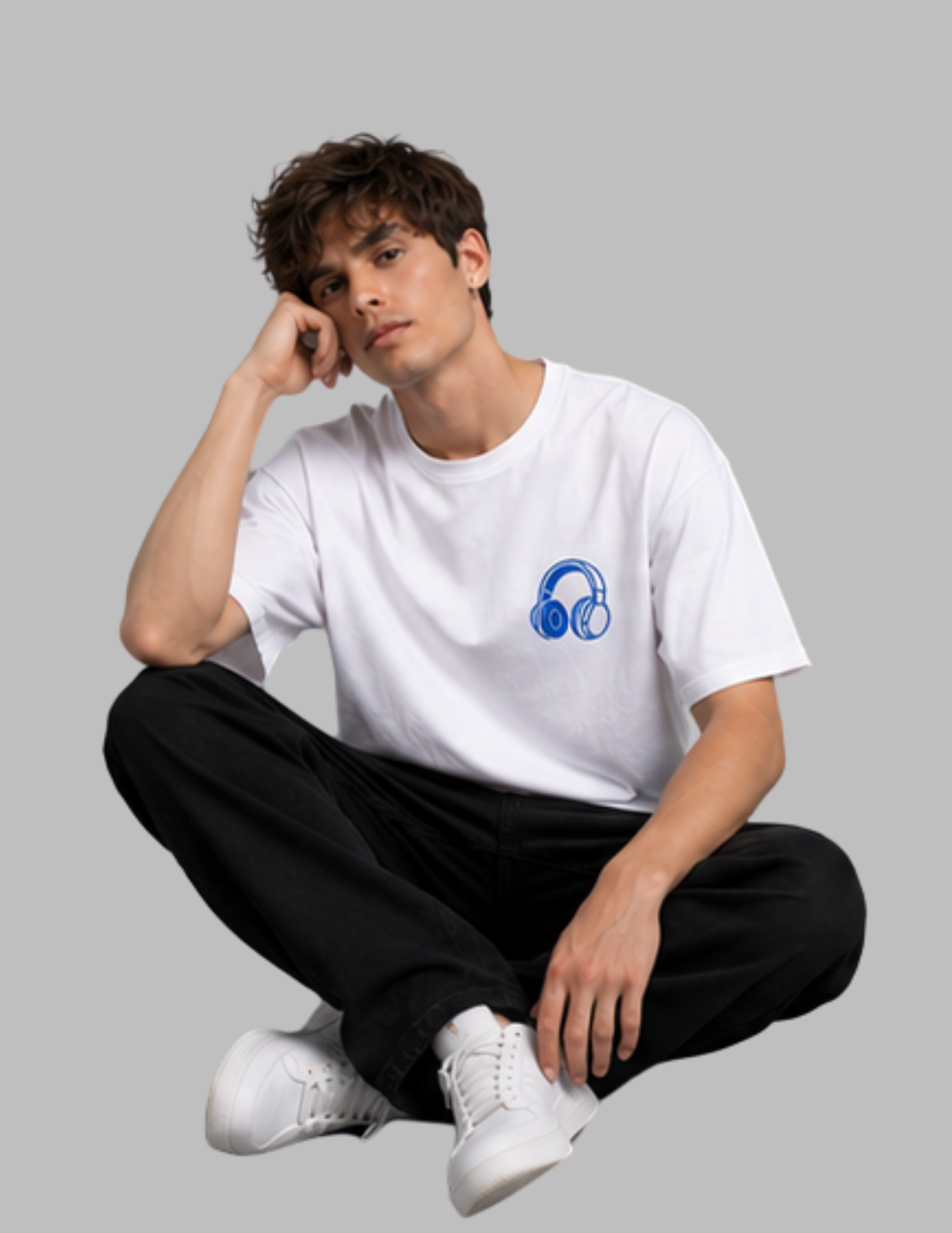 A young man sitting cross-legged against a solid gray background, wearing a white crewneck t-shirt with a blue headphones graphic on the left chest, paired with black pants and white sneakers.