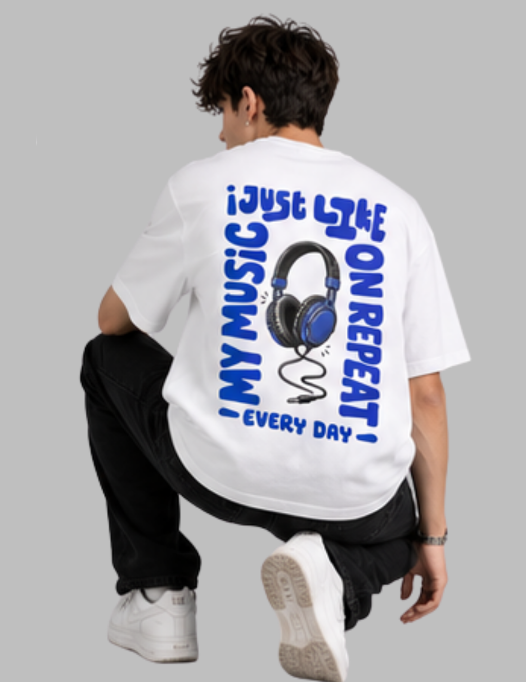 A man wearing a white t-shirt with a blue graphic print on the back, crouching in a studio setting against a solid gray background.