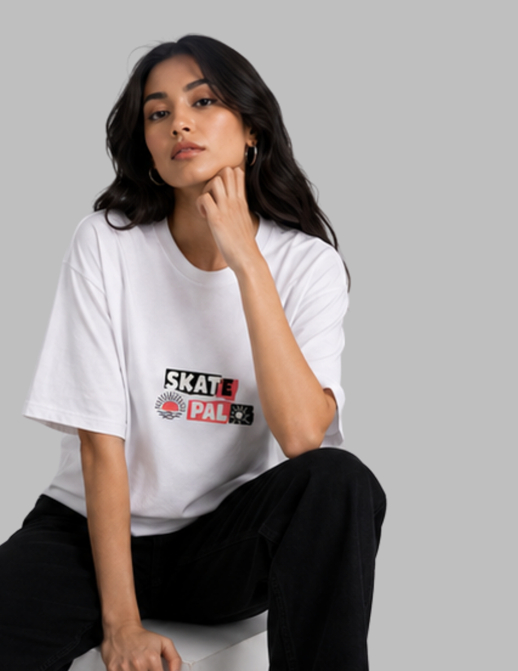 A woman wearing a white SkatePAL t-shirt with a colorful graphic logo on the chest, paired with black pants, posed against a solid gray studio background.
