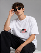 A man wearing a white short-sleeve t-shirt with a 'SKATE PA!' graphic on the chest, black sunglasses, and black pants, sitting against a gray studio background.