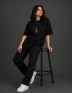 A woman wearing a black oversized t-shirt with a small brown graphic on the chest, black cargo pants, and white sneakers, sitting on a black stool against a dark gray studio background.