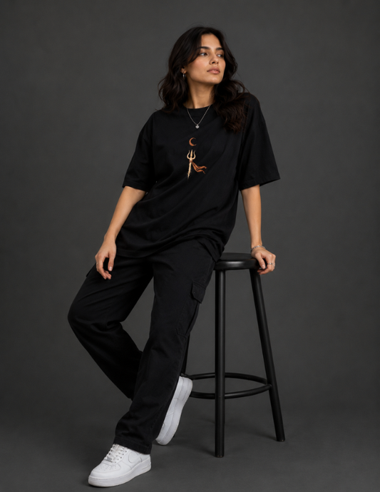 A woman wearing a black oversized t-shirt with a small brown graphic on the chest, black cargo pants, and white sneakers, sitting on a black stool against a dark gray studio background.