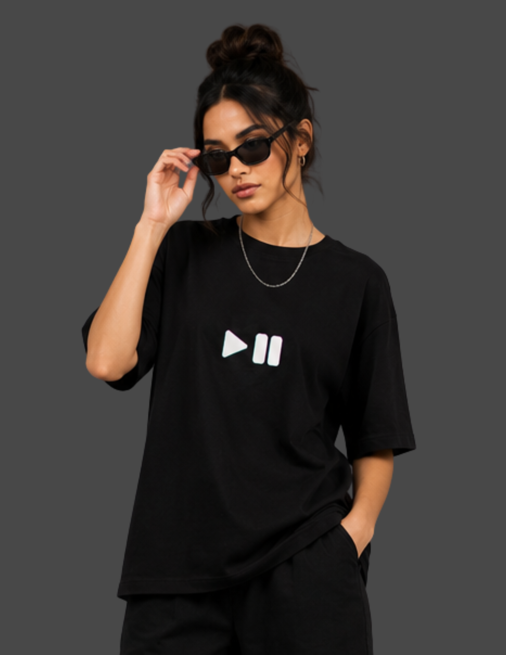 A woman modeling a black oversized t-shirt with a white play and pause icon printed on the chest, styled with black pants, sunglasses, and a silver necklace against a dark gray background.