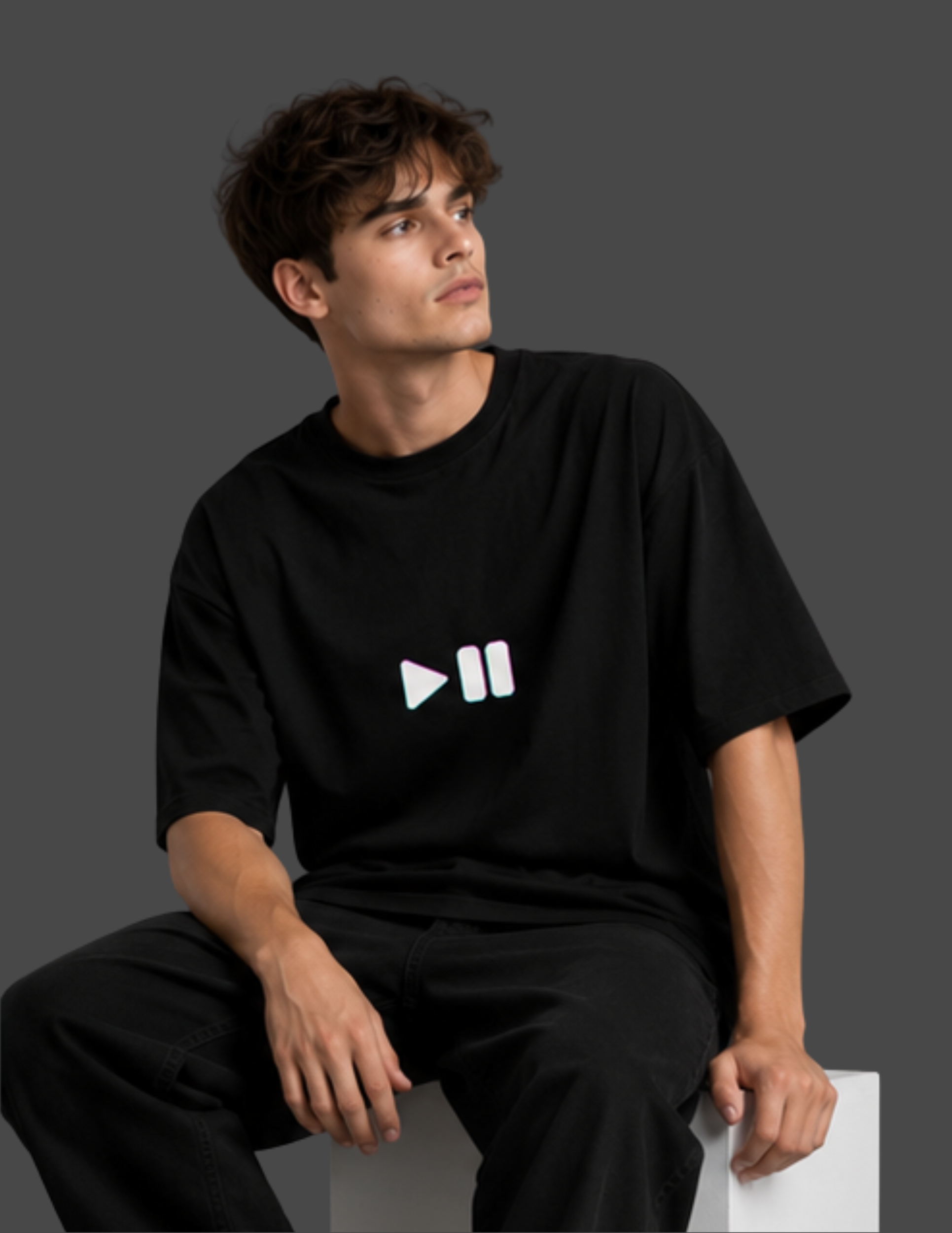 A man wearing a black oversized t-shirt with a white play and pause icon on the chest, sitting against a dark gray background.