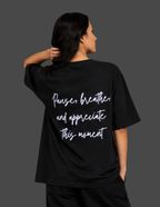 A woman shown from the back wearing a black oversized T-shirt with white script text that reads, 'Pause, breathe, and appreciate this moment.' The photo is shot against a dark gray studio background.