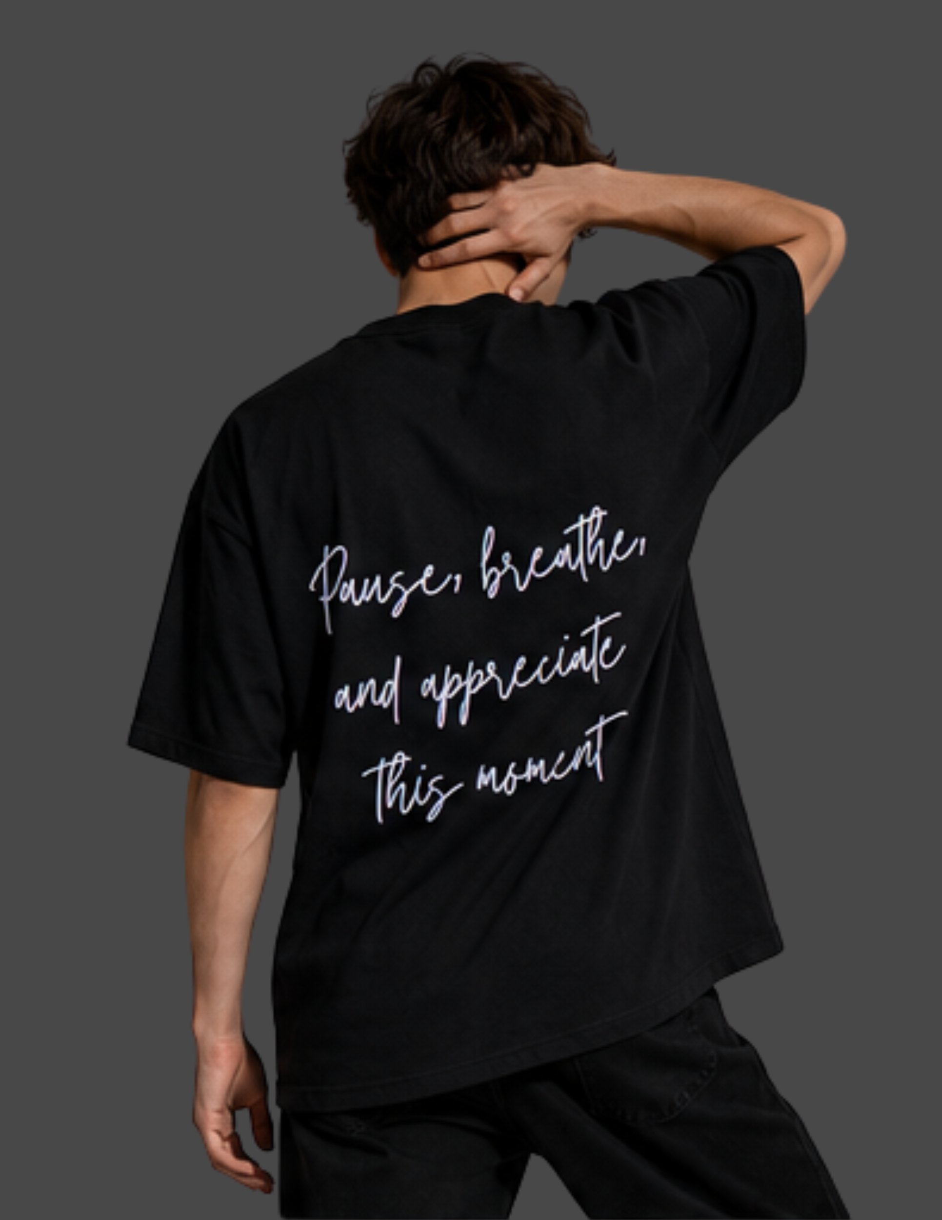 A man shown from behind wearing a black oversized t-shirt with white script text on the back that reads 'Pause, breathe, and appreciate this moment'.