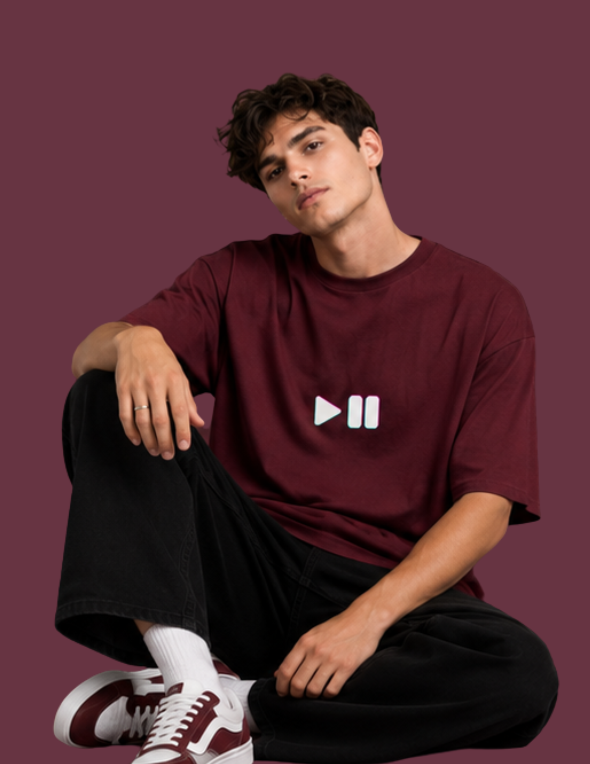 A young man sitting cross-legged against a solid dark burgundy background, wearing an oversized maroon t-shirt with a white play and pause icon on the chest, black jeans, white socks, and maroon and white sneakers.