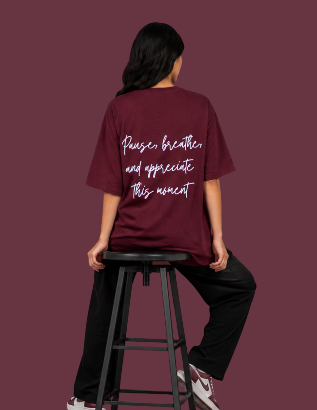 A person seated on a black stool, shown from the back, wearing an oversized burgundy T-shirt with white script text reading 'Pause, breathe and appreciate this moment' on the back. The background is a solid burgundy color.