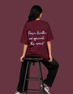 A person seated on a black stool, shown from the back, wearing an oversized burgundy T-shirt with white script text reading 'Pause, breathe and appreciate this moment' on the back. The background is a solid burgundy color.