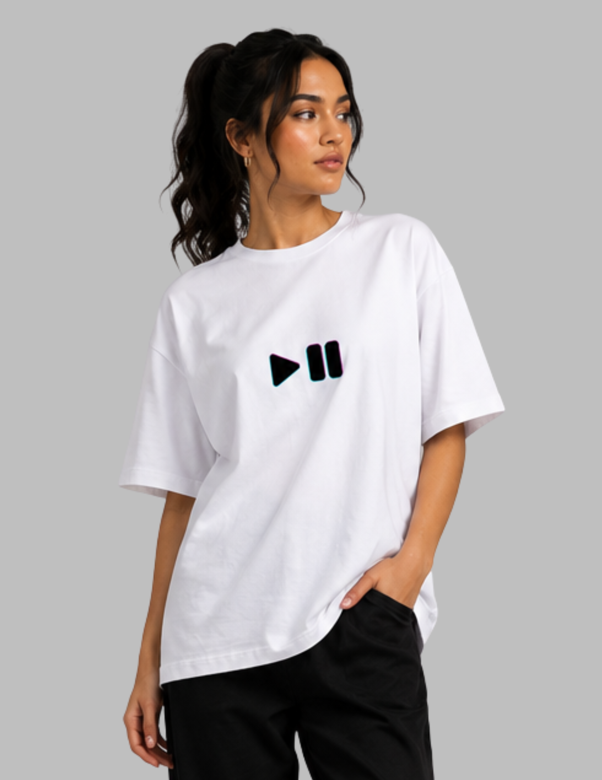 A woman wearing a white oversized t-shirt with a black play and pause icon printed on the chest, paired with black pants, set against a solid gray background.