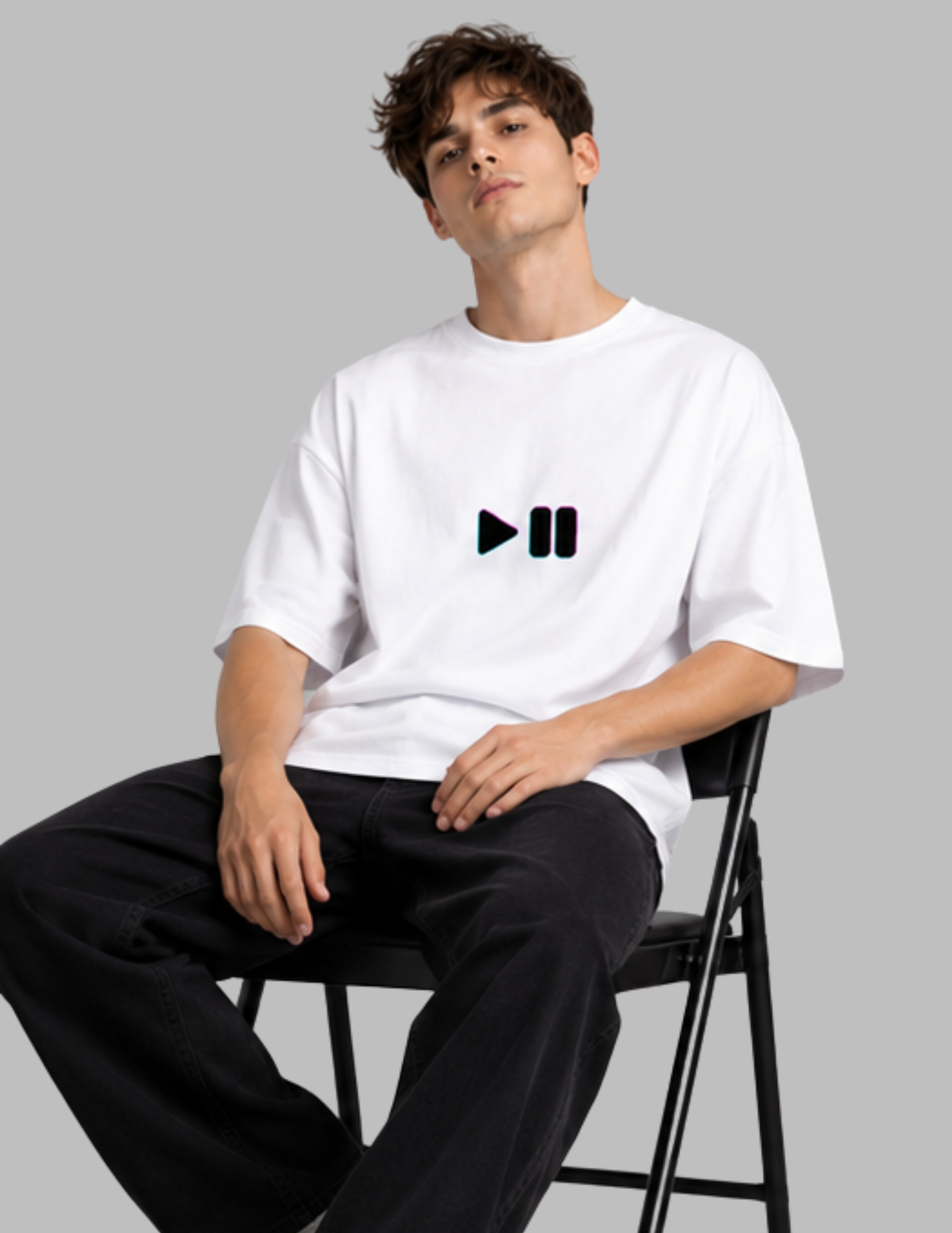 A man wearing a white oversized t-shirt with a black pause button graphic on the chest, paired with black pants, sitting on a black folding chair against a solid gray background.