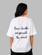 A woman viewed from the back wearing a white oversized T-shirt with dark navy script text that reads, 'Pause, breathe, and appreciate this moment.' The photo is shot against a plain light gray studio background.