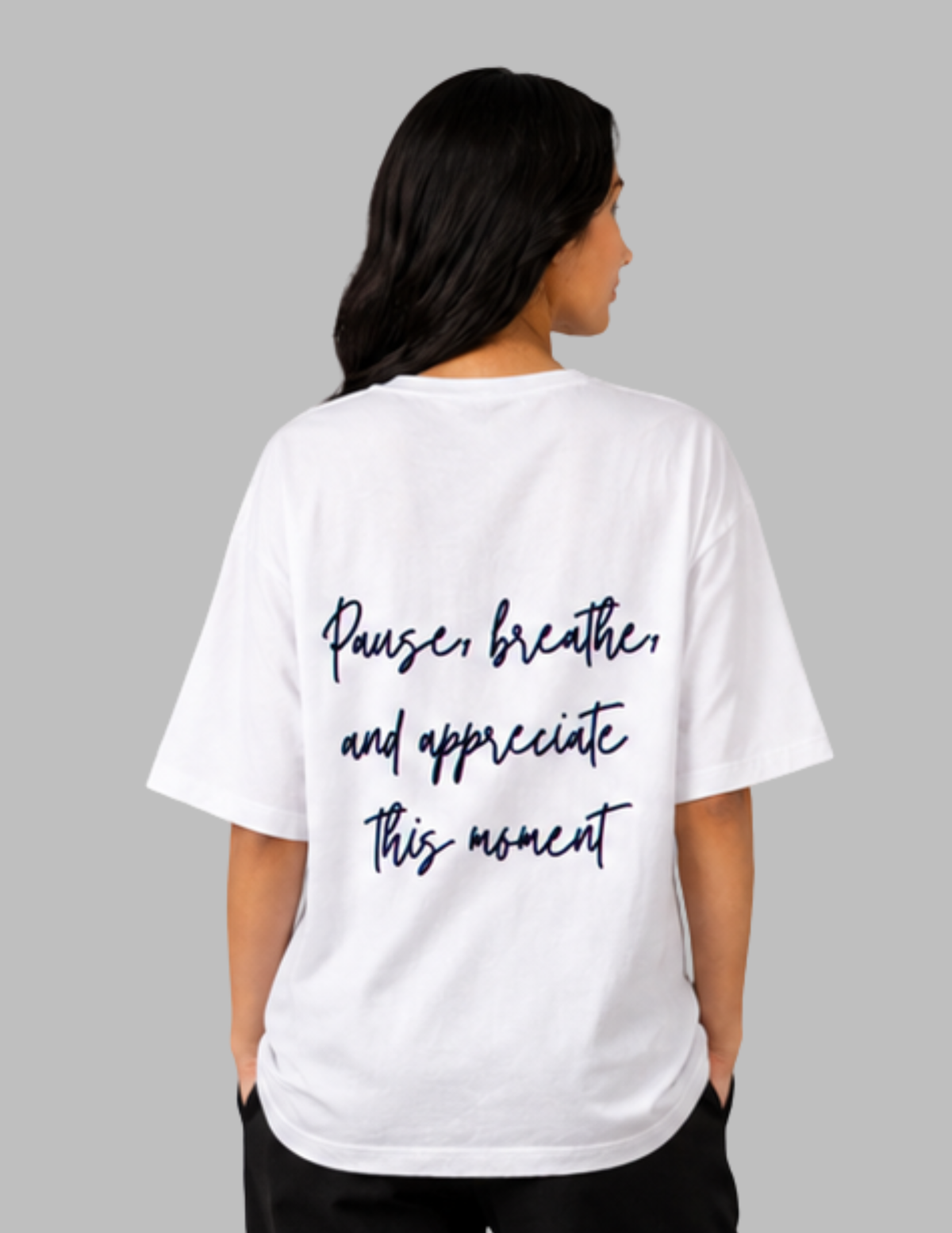 A woman viewed from the back wearing a white oversized T-shirt with dark navy script text that reads, 'Pause, breathe, and appreciate this moment.' The photo is shot against a plain light gray studio background.