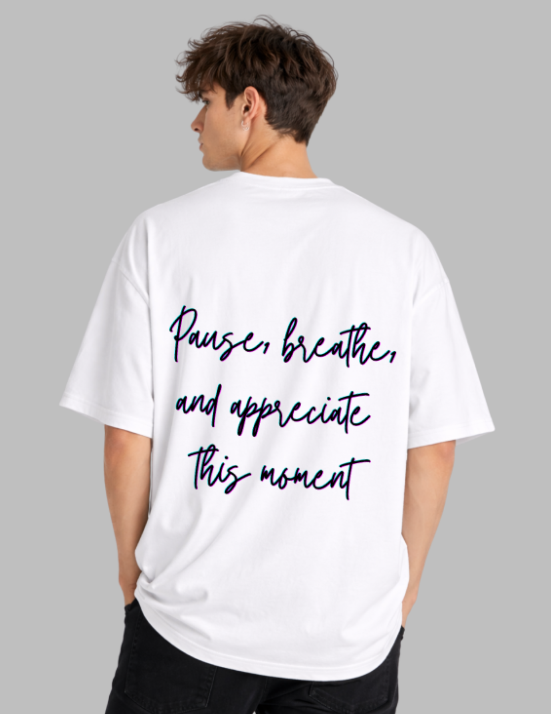 A man shown from the back wearing a white oversized T-shirt with the phrase 'Pause, breathe, and appreciate this moment' printed in dark navy script across the back, against a solid gray background.