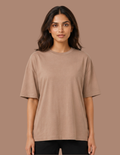 A woman modeling an oversized short-sleeve crewneck t-shirt in a solid taupe-brown color, paired with black pants, against a solid taupe background.