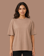 A woman modeling an oversized short-sleeve crewneck t-shirt in a solid taupe-brown color, paired with black pants, against a solid taupe background.