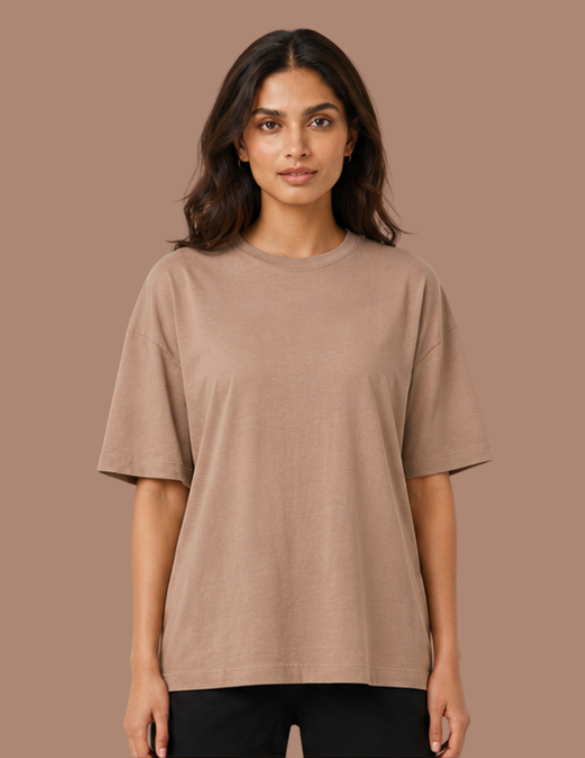 A woman modeling an oversized short-sleeve crewneck t-shirt in a solid taupe-brown color, paired with black pants, against a solid taupe background.