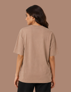 A woman shown from the back wearing a plain taupe short-sleeve t-shirt and black pants, set against a solid taupe background.