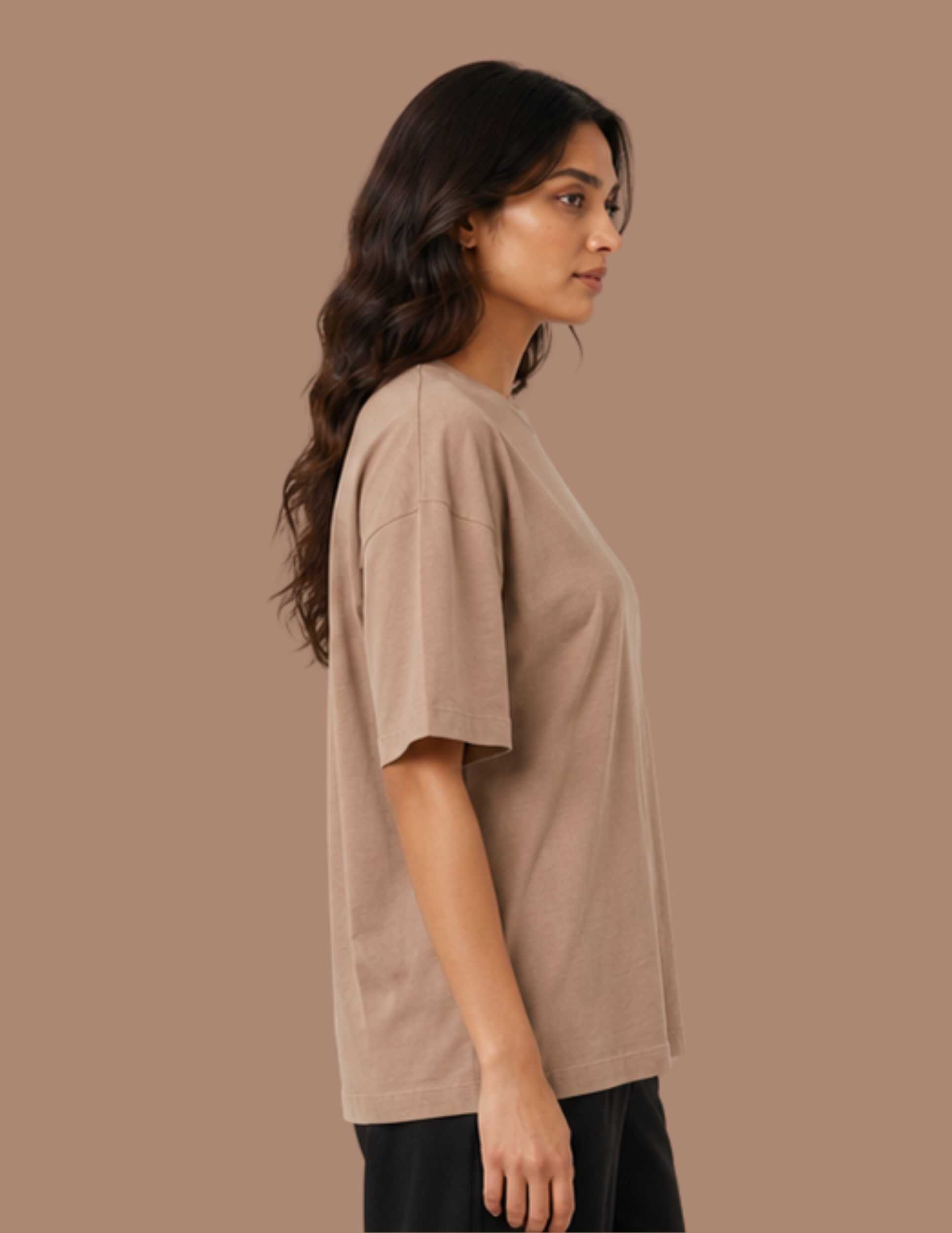 A woman shown in side profile wearing an oversized short-sleeve crewneck t-shirt in a warm taupe-beige color, paired with black pants, against a solid taupe background.