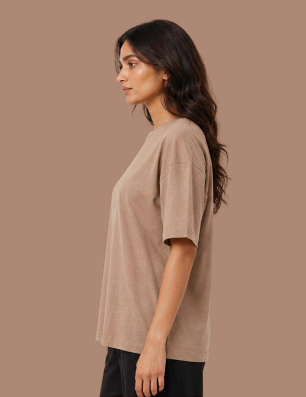 A woman shown in side profile wearing a loose, short-sleeve crew-neck t-shirt in a warm taupe-beige color, paired with black pants, against a solid taupe background.