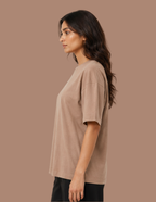 A woman shown in side profile wearing a loose, short-sleeve crew-neck t-shirt in a warm taupe-beige color, paired with black pants, against a solid taupe background.