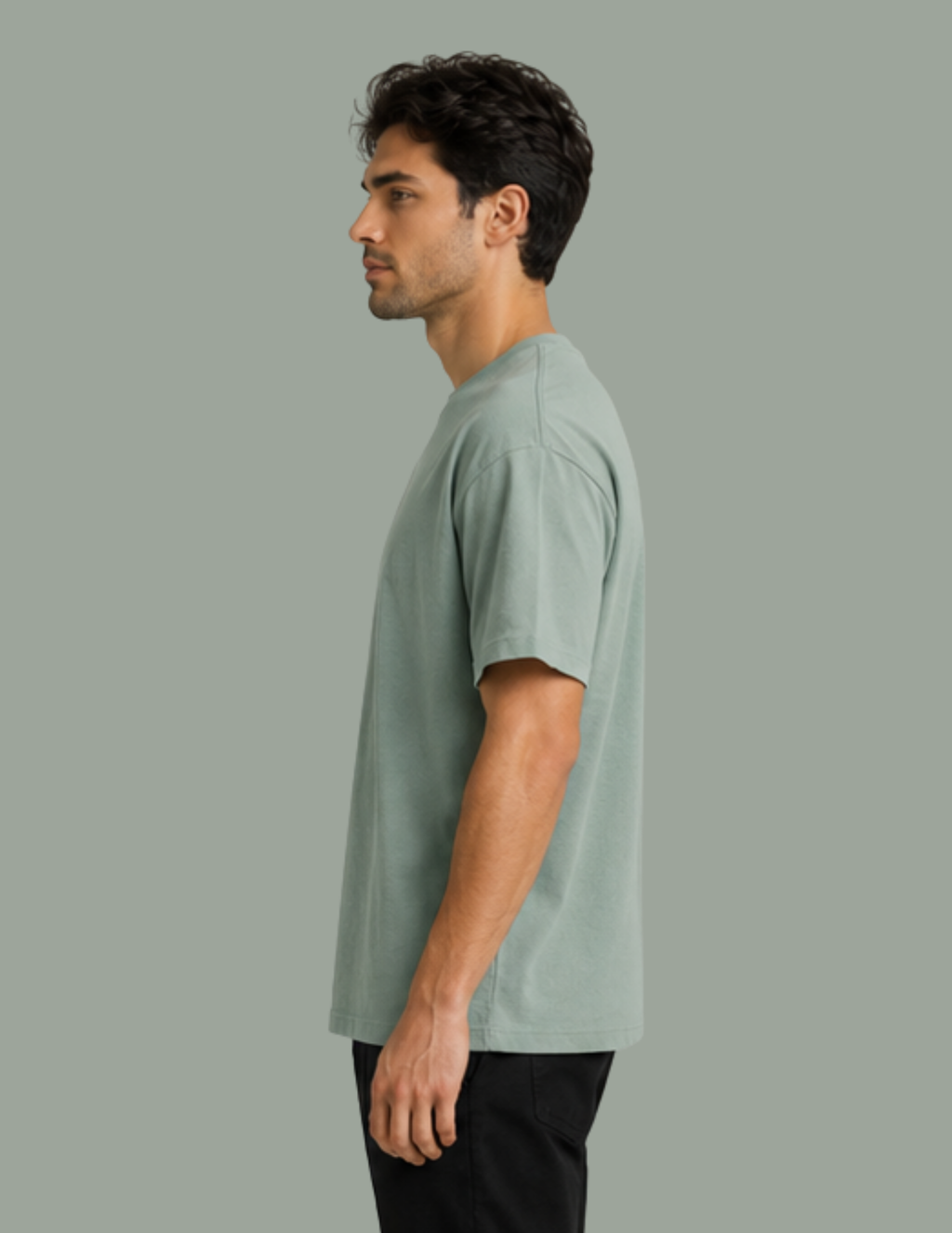 A man in profile wearing a light sage green short-sleeve crewneck t-shirt and black pants, standing against a solid sage green background.