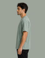 A man in profile wearing a light sage green short-sleeve crewneck t-shirt and black pants, standing against a solid sage green background.