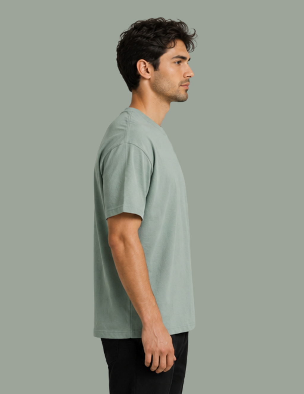 A man in profile wearing a light sage green crew-neck t-shirt and black pants, standing against a solid sage green background.