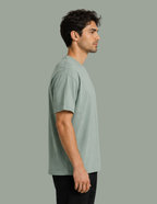 A man in profile wearing a light sage green crew-neck t-shirt and black pants, standing against a solid sage green background.
