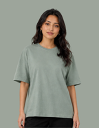 A woman modeling a short-sleeve crewneck t-shirt in a muted sage green color, paired with black pants, against a solid sage green background.