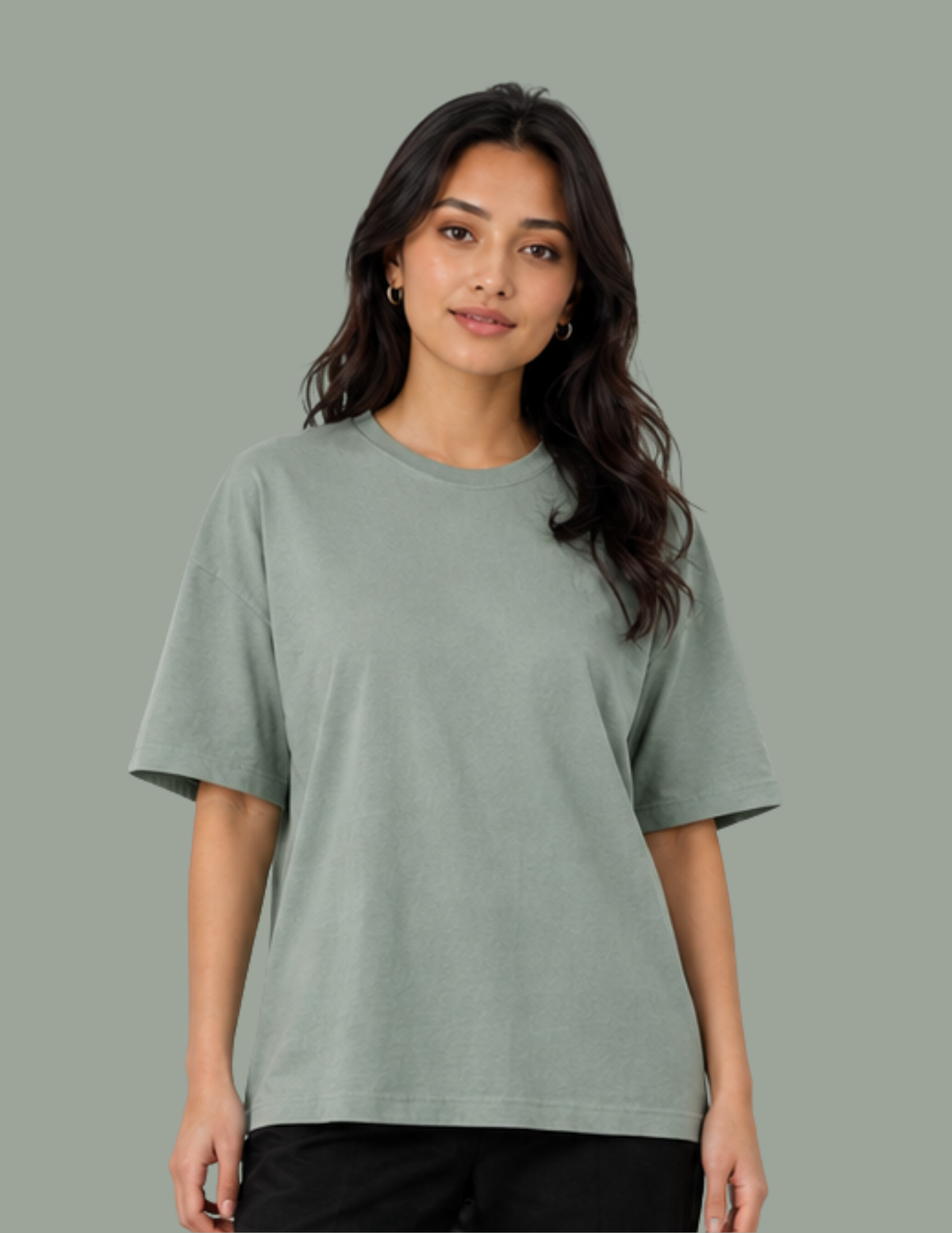 A woman modeling a short-sleeve crewneck t-shirt in a muted sage green color, paired with black pants, against a solid sage green background.