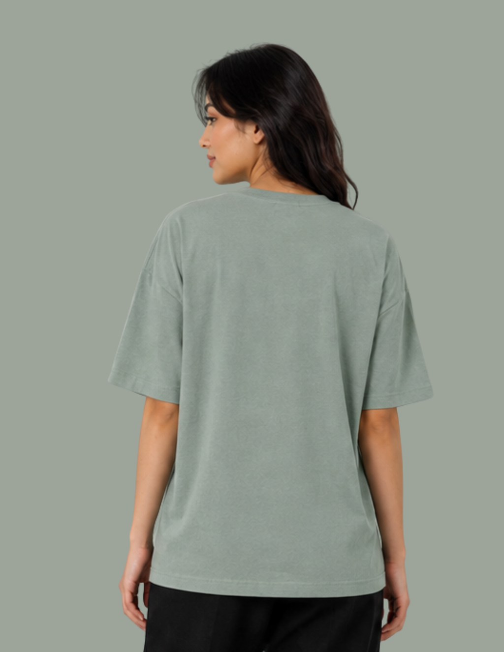 A woman shown from the back wearing an oversized sage green t-shirt and black pants, set against a solid sage green studio background.