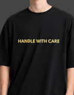 Black t-shirt with 'HANDLE WITH CARE' text in gold on a plain background