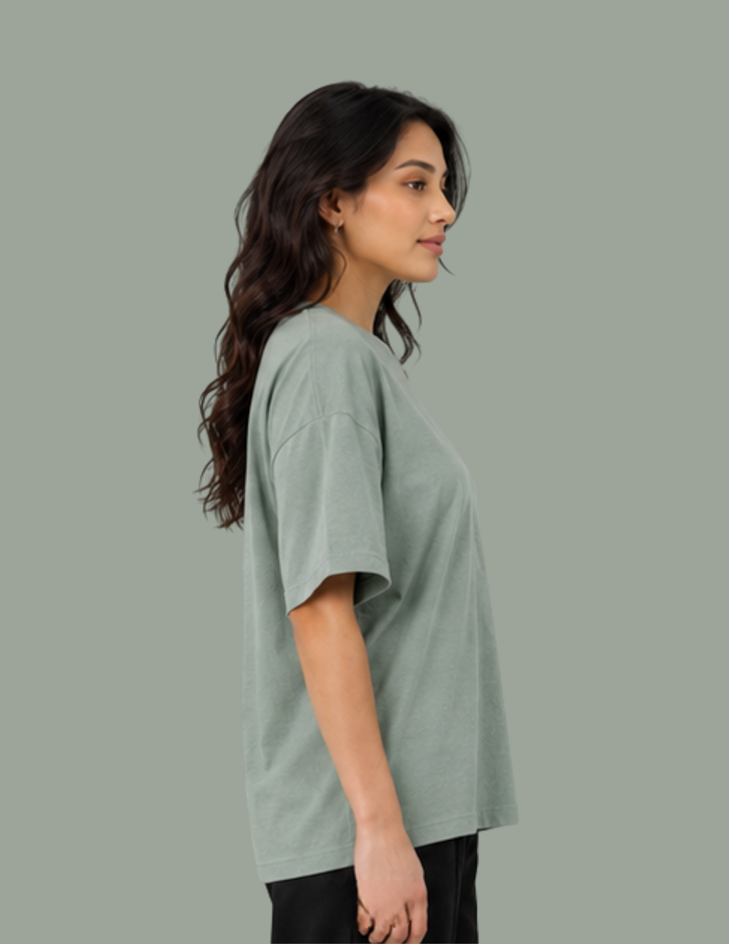 A woman shown in side profile wearing an oversized short-sleeve crewneck t-shirt in a muted sage green color, paired with black pants, against a solid sage green background.