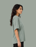 A woman shown in side profile wearing an oversized short-sleeve crewneck t-shirt in a muted sage green color, paired with black pants, against a solid sage green background.